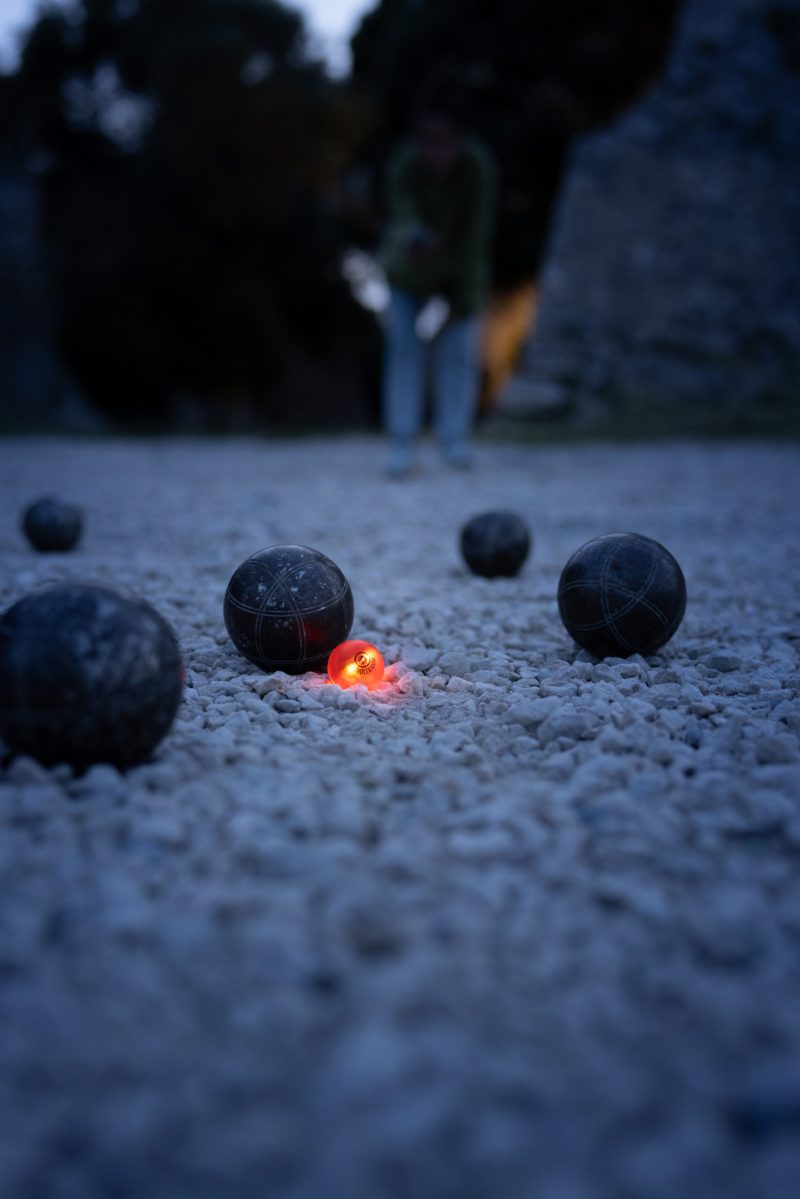 Cochonnet lumineux Cocholed orange au centre d'une partie de pétanque de nuit, entouré de boules noires