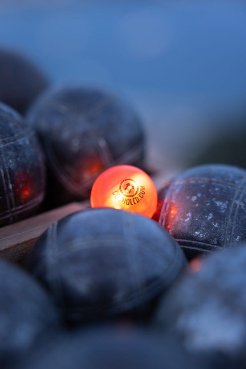 Cochonnet lumineux Cocholed orange parmi des boules de pétanque noires, éclairé en pleine partie de pétanque en extérieur lors du crépuscule