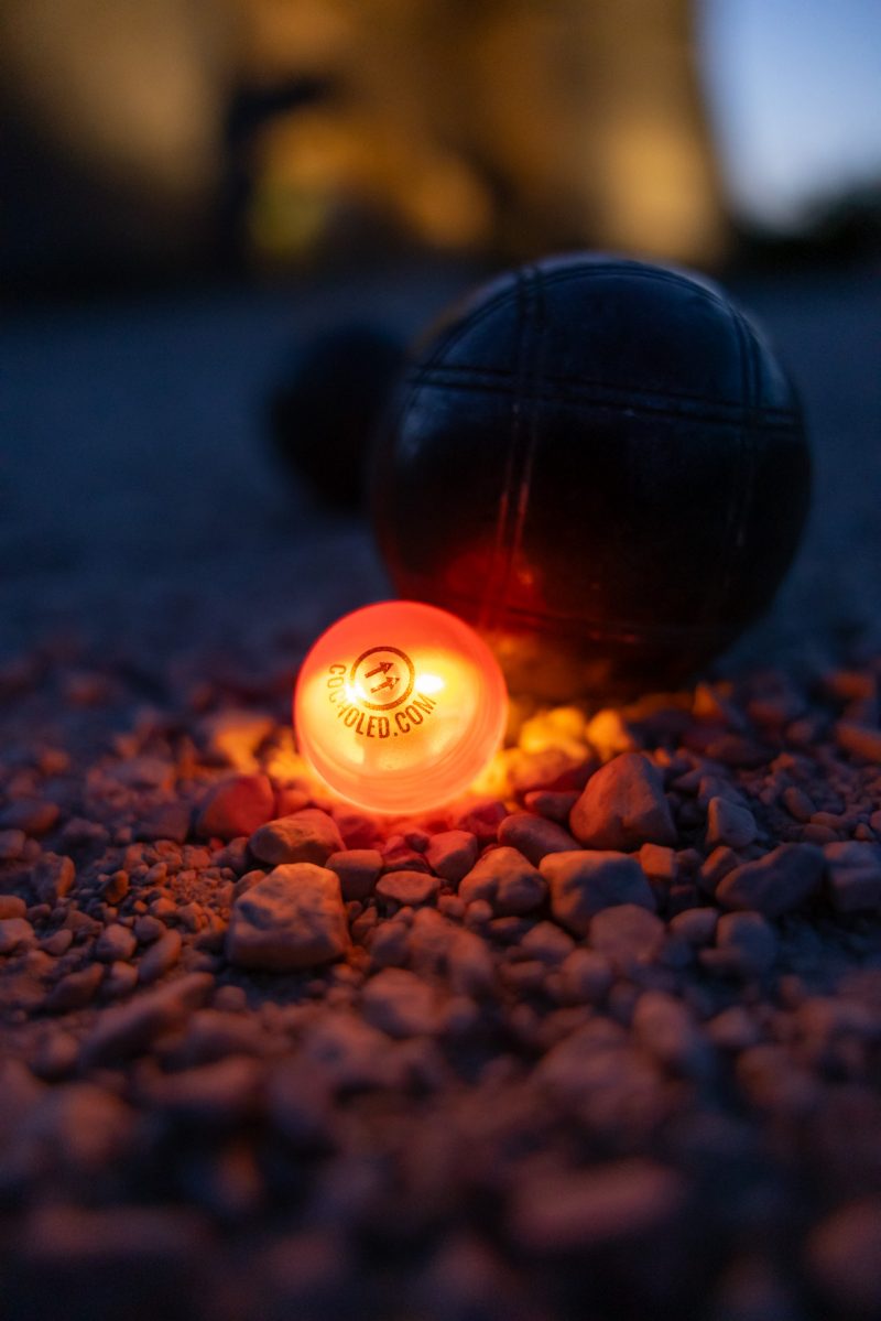 Cochonnet lumineux Cocholed orange posé sur le gravier, entouré de boules de pétanque, utilisé dans une partie de pétanque en extérieur lors de la tombée de la nuit